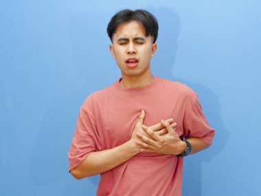 Sağlıksız Asyalı bir adam mavi arka planda acı ve üzüntüyle göğsüne dokunuyor..