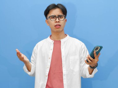 Elinde şok olmuş bir ifadeyle cep telefonu tutan ciddi bir Asyalı adamın portresi, mavi bir arkaplanda izole edilmiş, teknoloji veya reaksiyon temalı reklamlar için mükemmel..