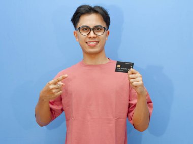 Asyalı adam mavi arka planda mutlu ve heyecanlı bir ifadeyle kredi kartını işaret ediyor. Finansal reklamlar ve terfiler için mükemmel..