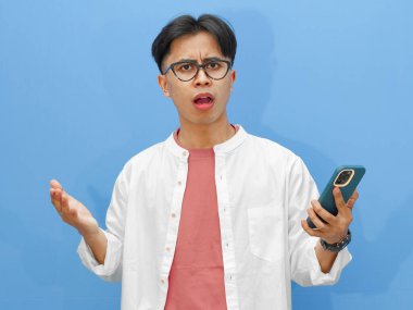 Elinde şok olmuş bir ifadeyle cep telefonu tutan ciddi bir Asyalı adamın portresi, mavi bir arkaplanda izole edilmiş, teknoloji veya reaksiyon temalı reklamlar için mükemmel..