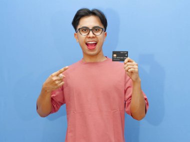 Asyalı adam mavi arka planda mutlu ve heyecanlı bir ifadeyle kredi kartını işaret ediyor. Finansal reklamlar ve terfiler için mükemmel..