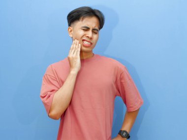 Acılı bir diş ağrısından muzdarip genç bir Asyalı üzgün bir ifadeyle yanağını tutuyor, mavi bir arka planda soyutlanmış. Diş ve ağız sağlığı konsepti.