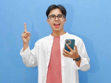 Elinde cep telefonu olan heyecanlı Asyalı bir adamın portresi mavi bir arka planda izole edilmiş neşeli bir ifadeyle işaret ediyor..