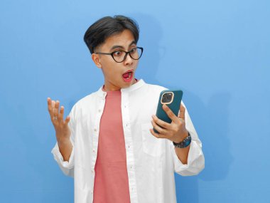 Cep telefonu kullanan Asyalı bir adamın portresi komik, şok olmuş bir ifadeyle, mavi arka planda izole edilmiş, reklam ve promosyon kullanımı için mükemmel..