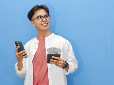 Cüzdanı para dolu Asyalı bir adamın portresi, cep telefonu kullanıyor, gülümsüyor ve mavi bir arka planda dikkatlice yukarı bakıyor..