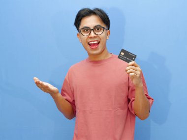 Asyalı heyecanlı genç adam elinde sürpriz bir ifadeyle kredi kartı tutuyor, mavi arka planda tek başına duruyor. Finans, bankacılık ve e-ticaret temaları için ideal.