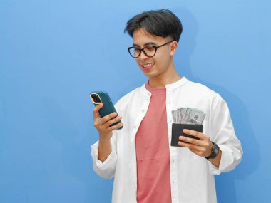 Cüzdanı para dolu gülümseyen Asyalı bir adamın portresi mavi arka planda cep telefonuna odaklanırken.