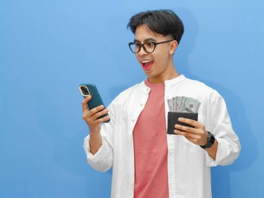 Cüzdanı para dolu mutlu bir Asyalı adamın portresi ve izole edilmiş mavi bir arka planda hayret verici bir ifadeyle cep telefonu kullanmak..