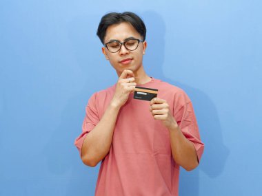 Elinde kredi kartı olan gülümseyen Asyalı genç bir adamın portresi, izole edilmiş mavi bir arka planda dururken düşünceli bir jest yapıyor..
