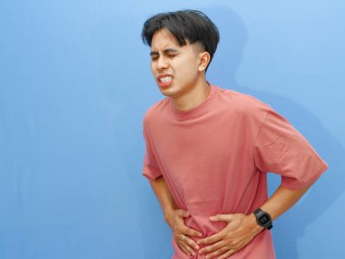 Sağlıksız bir adam midesine bastırıyor, acı çekiyor ve üzgün bir ifade sergiliyor, mavi arka planda izole edilmiş. Sağlık ve yaşam tarzı konularına uygun.