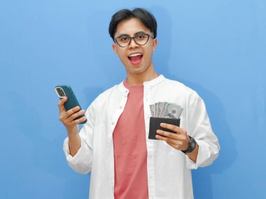 Cüzdanı para dolu mutlu bir Asyalı adamın portresi ve izole edilmiş mavi bir arka planda hayret verici bir ifadeyle cep telefonu kullanmak..