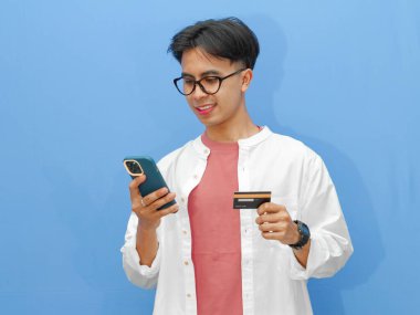 Elinde kredi kartı ve cep telefonu olan Asyalı genç bir adamın portresi, izole edilmiş mavi bir arka planda çevrimiçi ödeme yaparken gülümsüyor..