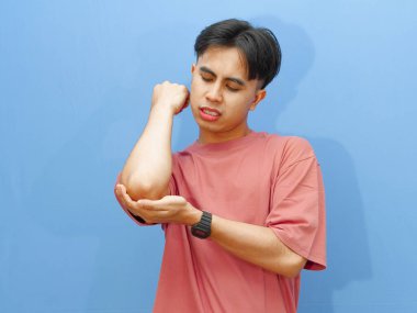 Dirseğini tutan genç, sağlıksız bir Asyalı adamın portresi, izole edilmiş mavi bir arka planda acı ve üzüntü gösteriyor..