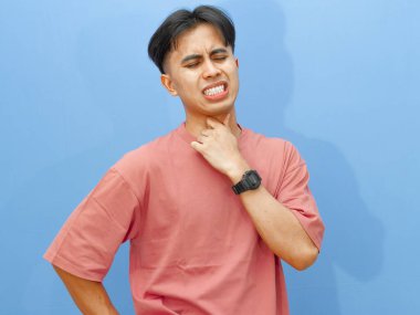 Hüzünlü ve acı dolu bir ifadeyle boğazını tutan sağlıksız bir gencin portresi, mavi bir arka planda izole edilmiş, rahatsızlık ve hastalık gösteriyor..