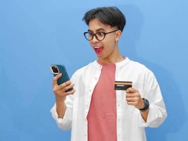 Kredi kartı tutan heyecanlı bir Asyalı adamın portresi, cep telefonunu online ödeme için kullanıyor, mavi arka plandaki başarıyı kutluyor..