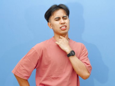 Hüzünlü ve acı dolu bir ifadeyle boğazını tutan sağlıksız bir gencin portresi, mavi bir arka planda izole edilmiş, rahatsızlık ve hastalık gösteriyor..