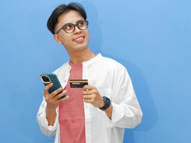 Kredi kartı ve para dolu cüzdanı olan mutlu bir Asyalı genç adam, izole edilmiş mavi arka planda bir düşünce hareketi yapıyor ve kafasını kaldırıyor..