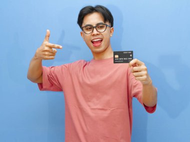 Asyalı adam mavi arka planda mutlu ve heyecanlı bir ifadeyle kredi kartını işaret ediyor. Finansal reklamlar ve terfiler için mükemmel..