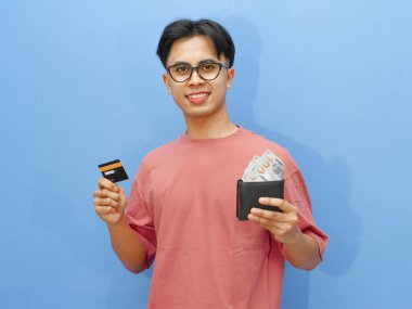 Asyalı bir adam para dolu bir cüzdan ve bir kredi kartı tutuyor. Güvenle gülümsüyor, mavi bir arka planda izole edilmiş..