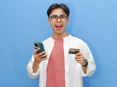 Kredi kartı tutan heyecanlı bir Asyalı adamın portresi, cep telefonunu online ödeme için kullanıyor, mavi arka plandaki başarıyı kutluyor..