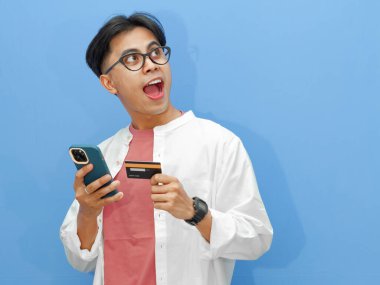 Kredi kartı tutan heyecanlı bir Asyalı adamın portresi, cep telefonunu online ödeme için kullanıyor, mavi arka plandaki başarıyı kutluyor..