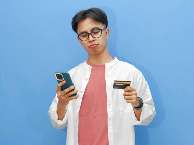 Kredi kartı tutan mutsuz bir Asyalı adamın portresi ve cep telefonu kullanması hüzünlü ve hayal kırıklığına uğramış bir ifadeyi mavi arka planda gösteriyor..