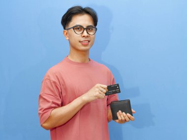 Asyalı özgüvenli bir adam cüzdanından banka kartı alıyor, gururla gülümsüyor, mavi arka planda soyutlanmış..