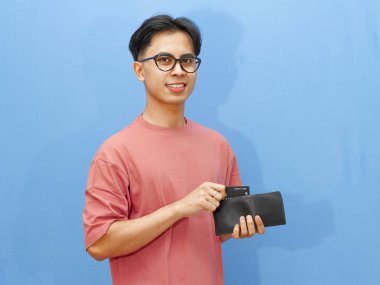 Asyalı özgüvenli bir adam cüzdanından banka kartı alıyor, gururla gülümsüyor, mavi arka planda soyutlanmış..
