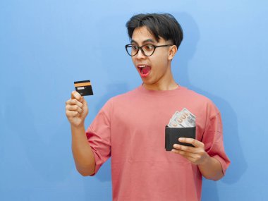 Elinde para dolu bir cüzdan ve kredi kartı olan Asyalı bir adam mavi arka planda komik ve heyecanlı bir ifade sergiliyor..