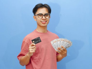 Portrait of an Asian man smiling while holding a credit card and cash, looking at the camera against a blue background. clipart