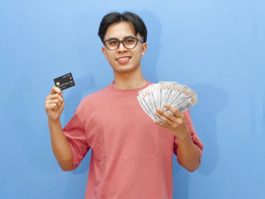 Elinde kredi kartı ve parayla gülümseyen Asyalı bir adamın portresi, mavi arka planda kameraya bakıyor..