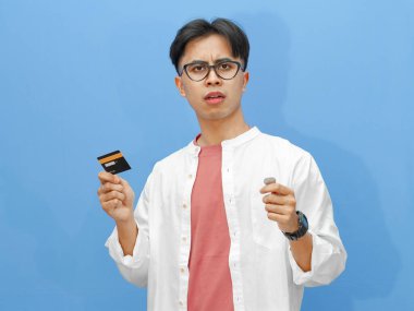 Elinde kredi kartı ve bozuk para tutan Asyalı genç bir adamın hüzünlü portresi izole edilmiş mavi bir arka planda mali hayal kırıklığını ifade ediyor..