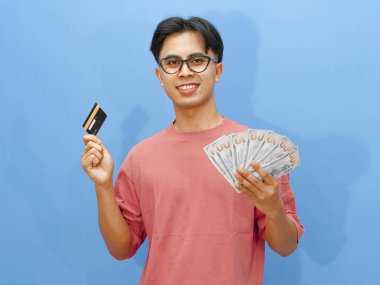 Elinde kredi kartı ve parayla gülümseyen Asyalı bir adamın portresi, mavi arka planda kameraya bakıyor..