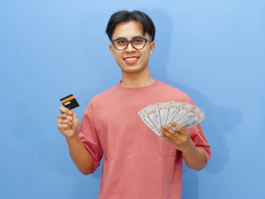 Elinde kredi kartı ve parayla gülümseyen Asyalı bir adamın portresi, mavi arka planda kameraya bakıyor..