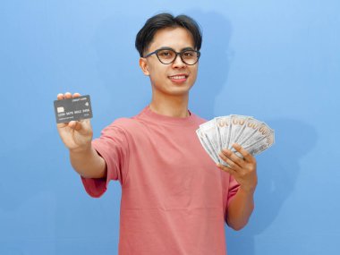 Portrait of an Asian man smiling while holding a credit card and cash, looking at the camera against a blue background. clipart