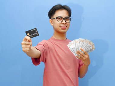 Elinde kredi kartı ve parayla gülümseyen Asyalı bir adamın portresi, mavi arka planda kameraya bakıyor..