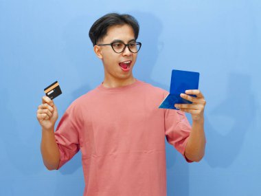 Asyalı heyecanlı adamın elinde banka kartı ve tasarruf defteri var. Şaşırtıcı bir ifadeyle, mavi arka planda izole edilmiş..