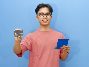 Kredi kartı ve banka tasarruf defteri tutan mutlu Asyalı adam, kameraya güvenle gülümsüyor, mavi arka planda izole edilmiş. Mali güvenlik ve bankacılık hizmetleri kavramı.