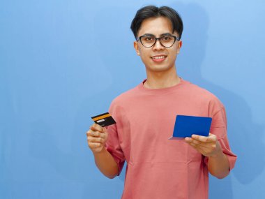 Kredi kartı ve banka tasarruf defteri tutan mutlu Asyalı adam, kameraya güvenle gülümsüyor, mavi arka planda izole edilmiş. Mali güvenlik ve bankacılık hizmetleri kavramı.