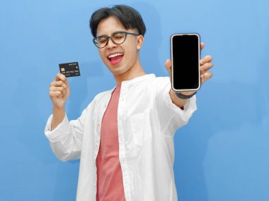 Sıradan kıyafetler içinde Asyalı bir adam mavi arka planda kredi kartı tutarken boş bir telefon ekranı gösteriyor. Bankacılık, teknoloji ve e-ticaret temaları için ideal..