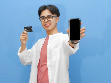Sıradan kıyafetler içinde Asyalı bir adam mavi arka planda kredi kartı tutarken boş bir telefon ekranı gösteriyor. Bankacılık, teknoloji ve e-ticaret temaları için ideal..