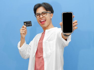 Sıradan kıyafetler içinde Asyalı bir adam mavi arka planda kredi kartı tutarken boş bir telefon ekranı gösteriyor. Bankacılık, teknoloji ve e-ticaret temaları için ideal..