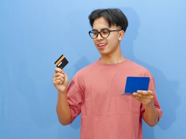 Elinde banka kartı ve tasarruf defteri tutan gülümseyen Asyalı bir adamın portresi, mavi arka planda izole edilmiş. Finans, bankacılık ya da ödeme kavramları için mükemmel.