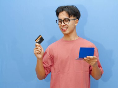 Elinde banka kartı ve tasarruf defteri tutan gülümseyen Asyalı bir adamın portresi, mavi arka planda izole edilmiş. Finans, bankacılık ya da ödeme kavramları için mükemmel.
