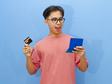 Asyalı heyecanlı adamın elinde banka kartı ve tasarruf defteri var. Şaşırtıcı bir ifadeyle, mavi arka planda izole edilmiş..