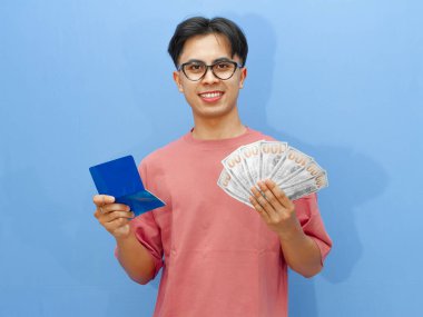 Portrait of a smiling Asian man holding money and a bank savings book, looking confidently at the camera on a blue background. clipart