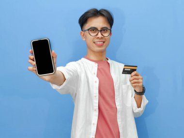 Sıradan kıyafetler içinde Asyalı bir adam mavi arka planda kredi kartı tutarken boş bir telefon ekranı gösteriyor. Bankacılık, teknoloji ve e-ticaret temaları için ideal..