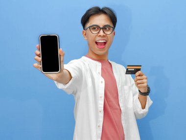 Sıradan kıyafetler içinde Asyalı bir adam mavi arka planda kredi kartı tutarken boş bir telefon ekranı gösteriyor. Bankacılık, teknoloji ve e-ticaret temaları için ideal..