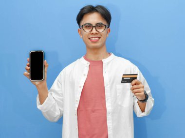 Sıradan kıyafetler içinde Asyalı bir adam mavi arka planda kredi kartı tutarken boş bir telefon ekranı gösteriyor. Bankacılık, teknoloji ve e-ticaret temaları için ideal..