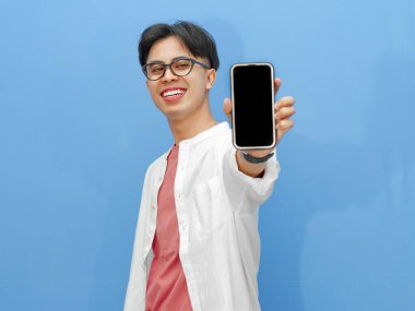 Sıradan giyinmiş genç bir Asyalı 'nın portresi mavi arka planda izole edilmiş boş bir cep telefonu ekranını işaret ediyor..